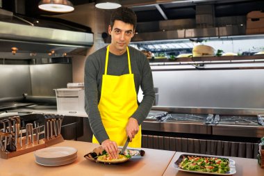 Smiling chef in small bistro prepares delicious burger in busy kitchen. clipart