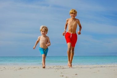 Two happy boys brothers big and small run on the sand beach smiling on summer vacation