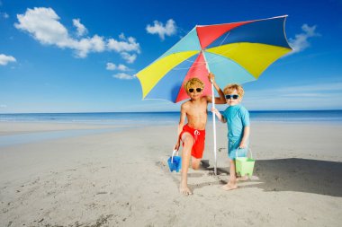 Two cute brother boys seat in sunshades play with sand buckets on the beach pose under parasol smiling over ocean on background