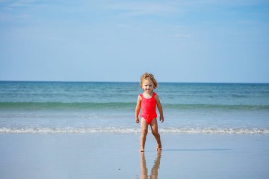 Little cute girl stand on a sand beach over ocean waves smiling at summer vacation