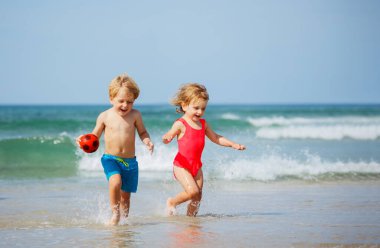 Two happy little children boy with sister girl run on the sand beach smiling on summer vacation holding ball