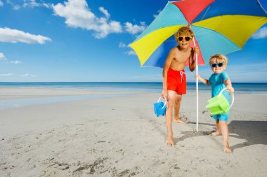 Two happy boys stand in sunshades play with sand buckets on the beach pose under parasol smiling over ocean on background