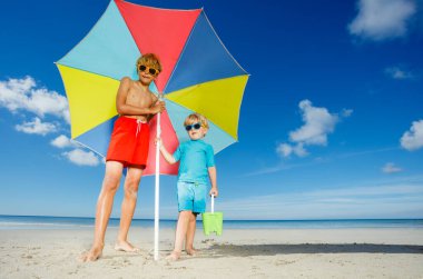 Portrait of two boys wear sunglasses stand on the beach under parasol smiling over ocean on background low angle image