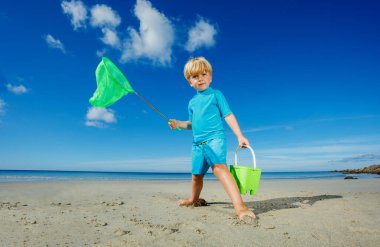 Little blond boy catcher of small critters crabs and shrimps on the sand beach during low tide holding bucket standing on the ocean shore