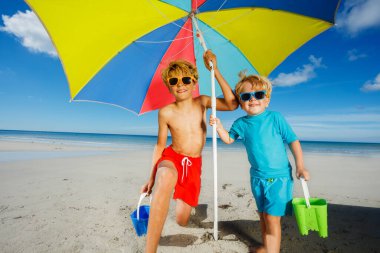 Two children in sunglasses under parasol on a sand ocean sun beach during summer vacation