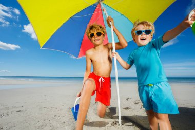 Portrait of two brother boys in sunshades pose on the beach under parasol smiling over ocean on background