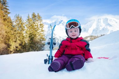 Little girl sit in snow with small mountain ski in sport outfit including helmet and skiing goggles over mountains
