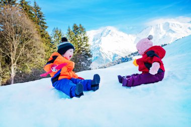 Two happy little kids boy and girl sit in snow throw snowballs at each other having fun