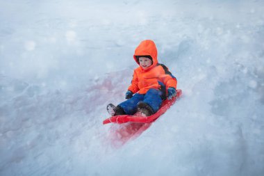 Happy young boy sled downhill on the sledge wearing sport winter outfit during snowfall