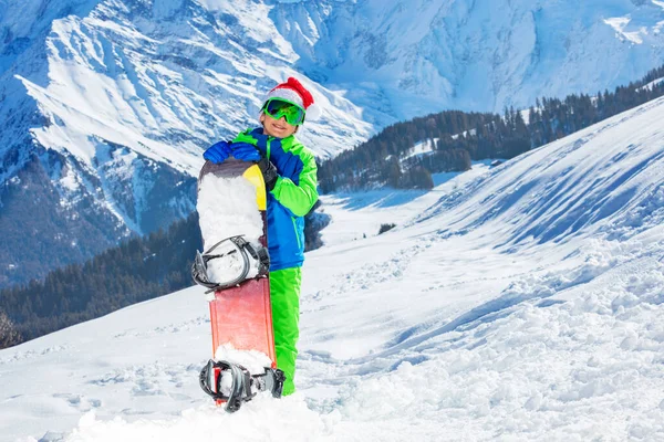 Boy in Santa Claus hat and ski googles stand with snowboard over the mountains on background