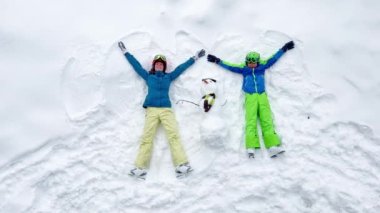 Ski vacation concept - two people: woman and young boy laying in the snow waving hands with snowman in winter outfit, helmets, top view from above