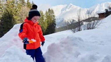 Little boy in orange coat play with shovel and snow outside on sunny winter day