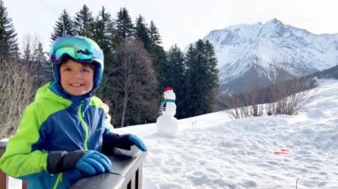 Happy boy on pose on the balcony smiling in ski outfit glasses and helmet with mountain peak on background