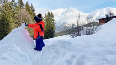Little boy in orange coat play with shovel and snow outside on sunny winter day