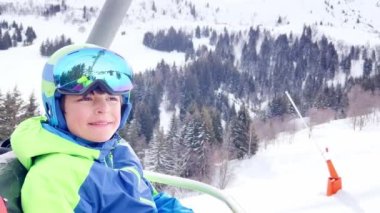 Close portrait of a boy sit in sport outfit with helmet mask and ski going up on chairlift