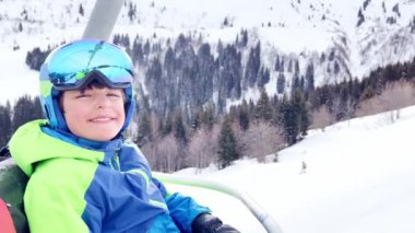 Close portrait of a boy sit in sport outfit with helmet mask and ski going up on chairlift