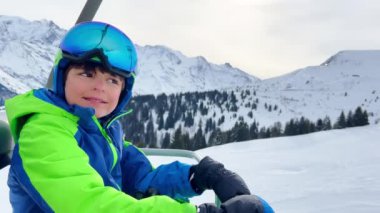 Close portrait of a boy sit in sport outfit with helmet mask and ski going up on chairlift