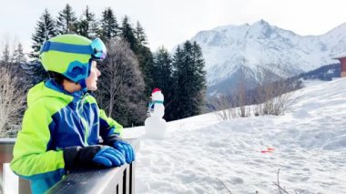 Happy boy on pose on the balcony smiling in ski outfit glasses and helmet with mountain peak on background