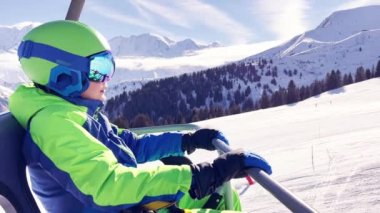 Close portrait of a boy sit in sport outfit with helmet mask and ski going up on chairlift