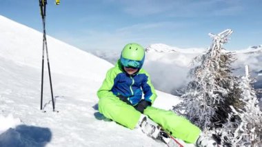 Little boy with alpine ski sit in snow observing the mountain over peaks covered by clouds on background