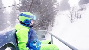 Close portrait of a boy sit in sport outfit with helmet mask and ski going up on chairlift