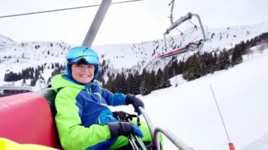 Close portrait of a boy sit in sport outfit with helmet mask and ski going up on chairlift