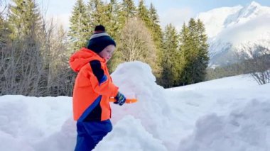 Little boy in orange coat play with shovel and snow outside on sunny winter day