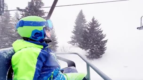 男の子の近い肖像画はヘルメットマスクとスキーがリフトに乗って行くスポーツ服に座っている — ストック動画