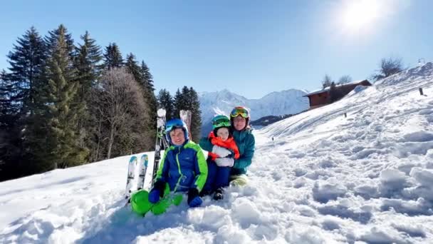 スキー休暇で楽しむ2人の若い男の子とスポーツ衣装の母親が山の上に一緒に座っています 家族の冬の休暇 — ストック動画
