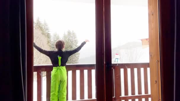 小男孩穿着冬季运动滑雪服走出阳台 兴高采烈地看着雪山森林 — 图库视频影像