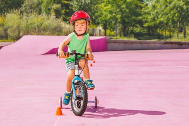 Cute little blond boy ride small bicycle with learning wheels around cones in the riding park