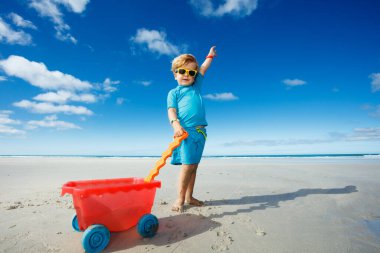 Happy little boy in sunglasses walk on the white sand beach playing with toy cart lifting hand up