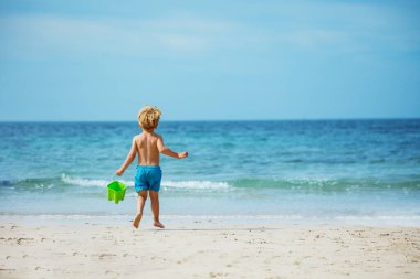 Little blond boy run to ocean waves on sand beach carry toy bucket with water view from behind