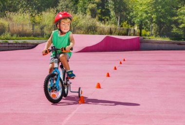 Little blond boy ride small bicycle with learning wheels roll around orange cones on color surface of the park