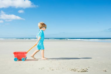Happy little boy in sunglasses walk on the white sand beach playing with toy cart side view