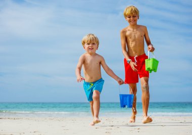 Two happy smiling boys run with water buckets from sea waves on sand beach during summer vacations