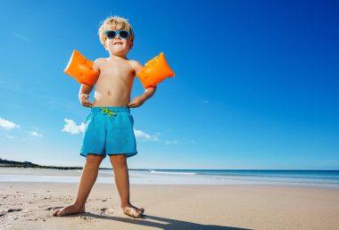Portrait of little blond child boy stand posing on the ocean beach with waves wearing inflatable shoulder straps