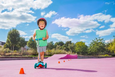 Little boy on the kick scooter ride around orange cones on the skate park with color surface in red helmet
