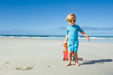 Happy cute boy in sunglasses stand on the white sand beach playing with toy cart