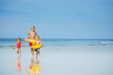 Group of three cute kids boys holding inflatable toy buoys doughnut and duck having fun running on the sand beach image with copy space