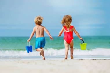 Little boy and a girl, siblings run barefoot to ocean waves sand beach carry toy buckets with water view from behind