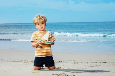 Cute handsome little blond boy sit on the sand hold toy boat playing at the sea beach during summer vacation