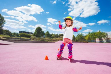 Smiling happy little girl learn to skate on rollers, stand rolling in helmet and protection at color skatepark surface