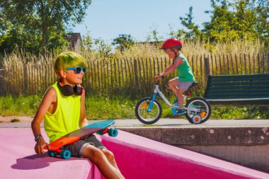 Cute preteen boy with green hair sit holing skate on a ramp at city skatepark wearing shades over little child on a bicycle
