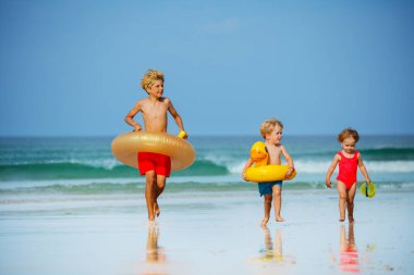 Three cute kids children with inflatable toy buoys doughnut and duck having fun running on the sand beach