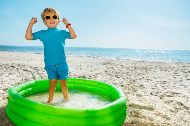 Cute blond little boy stand playing in inflatable pool in sunglasses lifting hands and smile