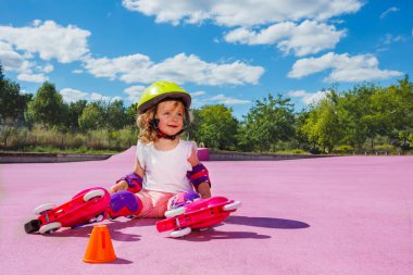 Cute little girl learn to skate on rollers, sit with helmet and protection on the color floor of the park