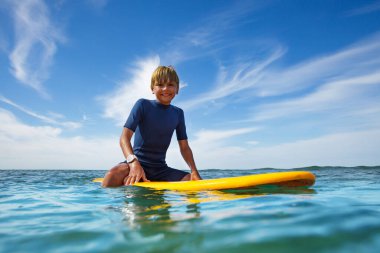 Smiling boy sit on the surf board looking at camera in ocean enjoying surfing school lesson
