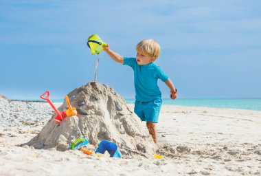 Cute little blond boy play with sand pouring water from plastic bucket on the sea beach during vacation
