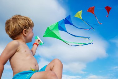 Close-up of a little boy sit holding many kites in hand and looking at them smiling over blue sky